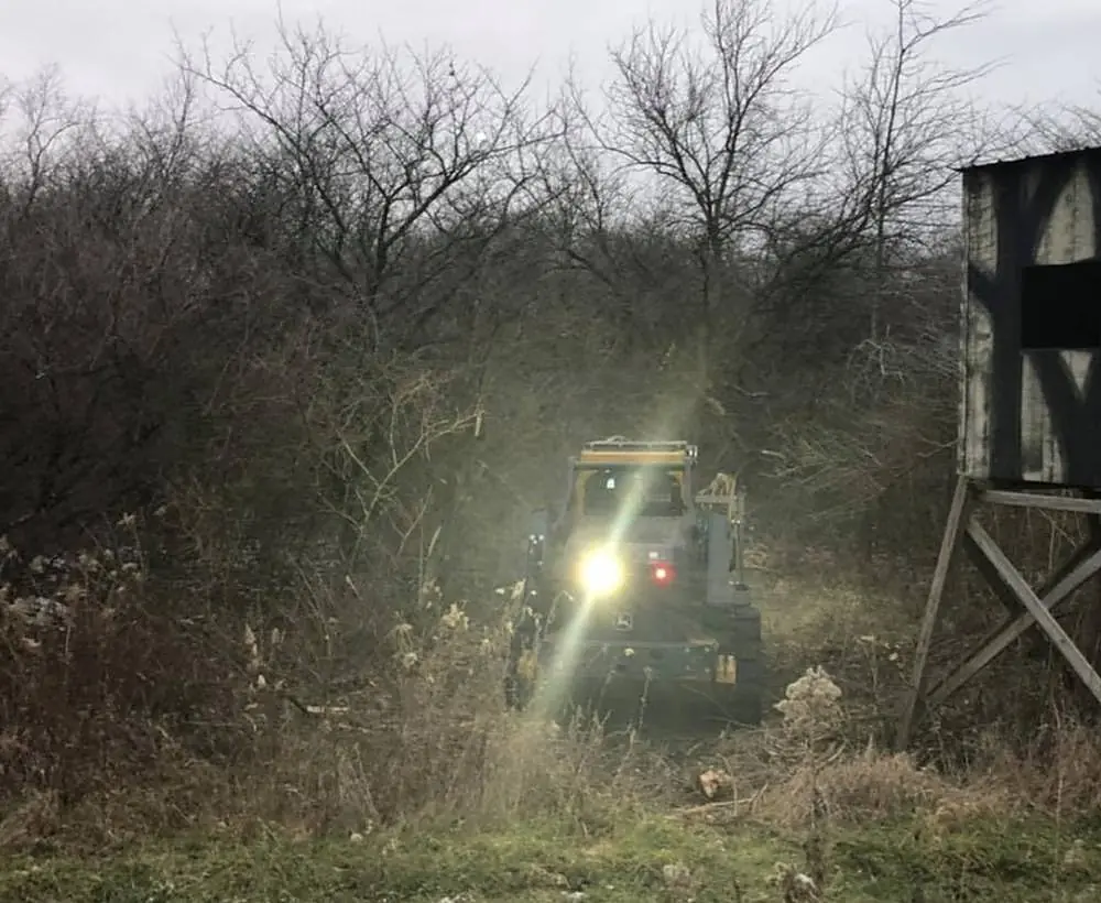 Tracked forestry mulcher working into heavy brush at dusk with its work lights on